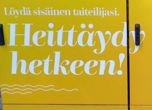 Kuva, jossa valkea teksti "Löydä sisäinen taiteilijasi. Heittäydy hetkeen!" keltaiseksi maalatussa sähkökaapissa kadulta.